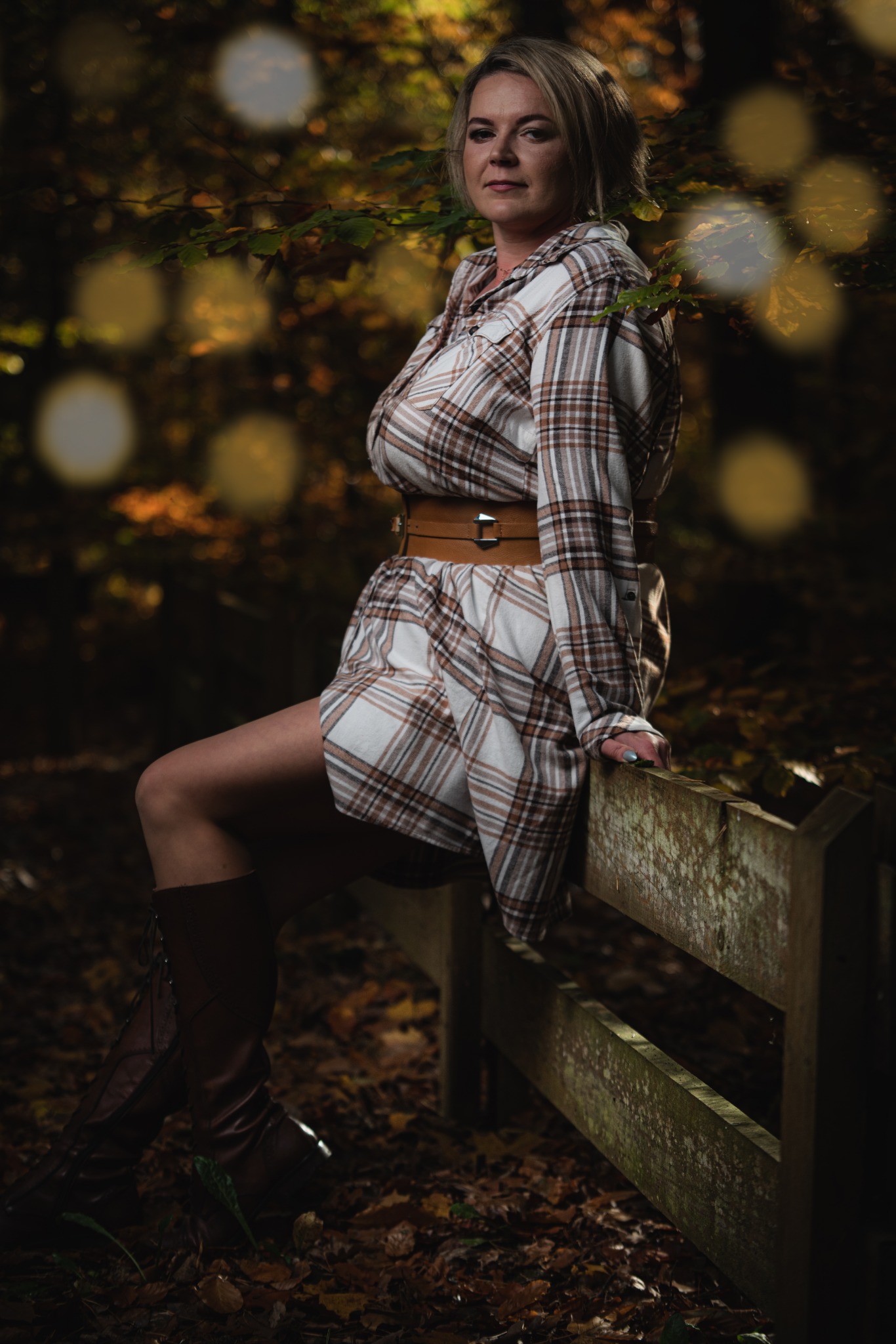 Kobieta w kraciastej sukience, skórzanym pasie i wysokich butach siedzi na drewnianym płocie na tle jesiennego lasu z bokeh.