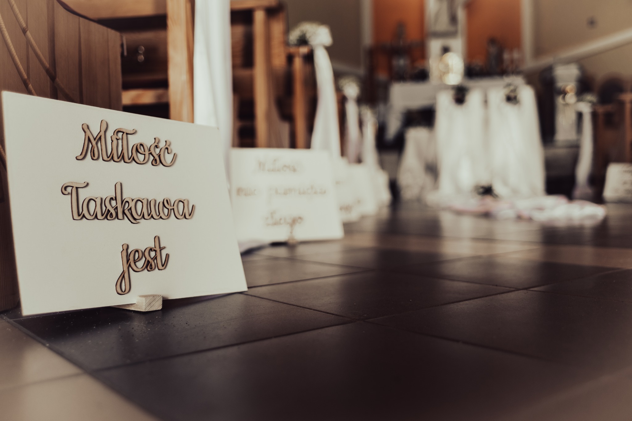 Dekoracja ślubna: tabliczka z napisem 'Miłość łaskawa jest' wykonana z drewnianych liter, ustawiona na podłodze, w tle dekoracje z białych tkanin.