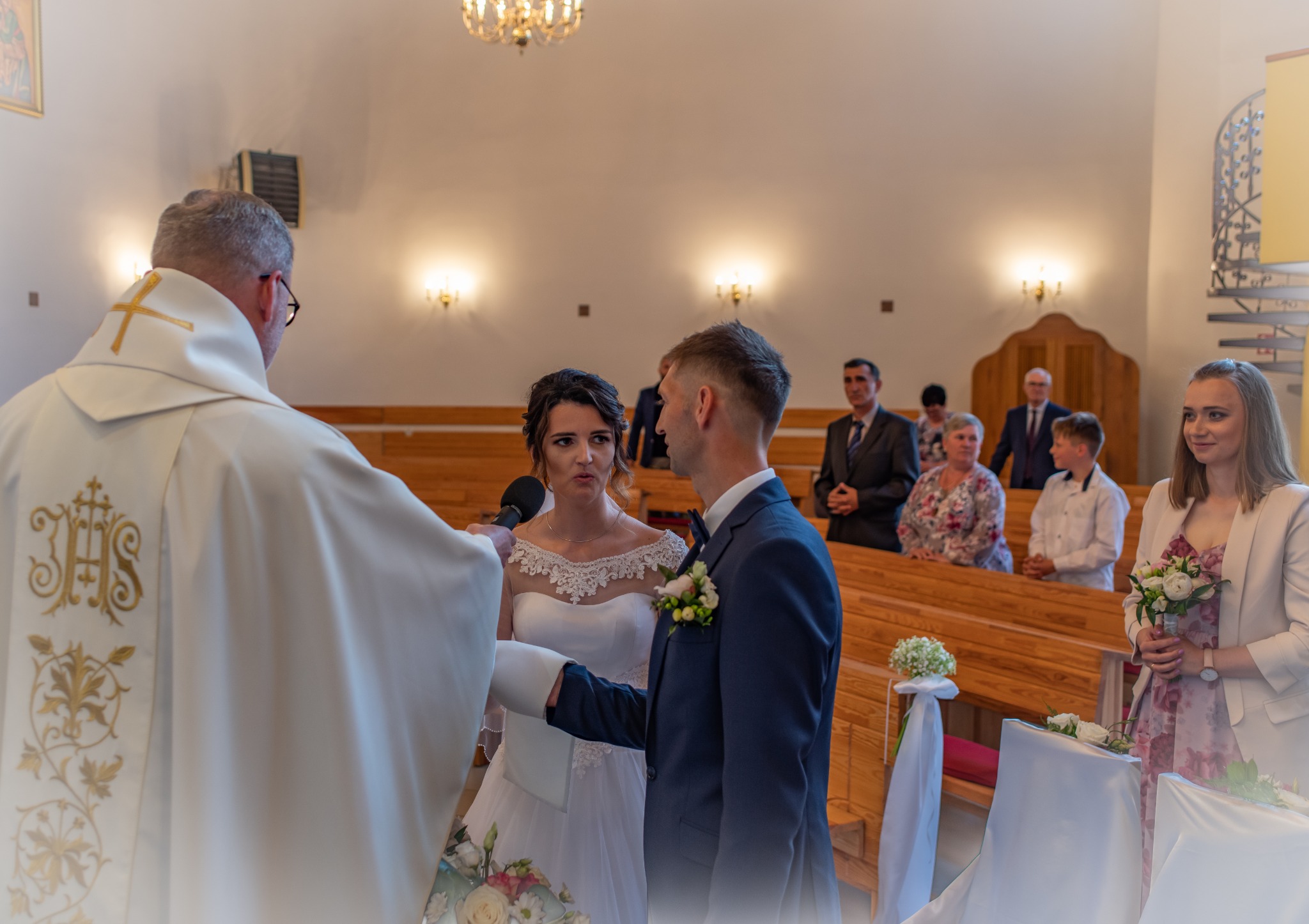 Para młoda podczas ceremonii ślubnej w kościele, ksiądz udziela błogosławieństwa, w tle goście.