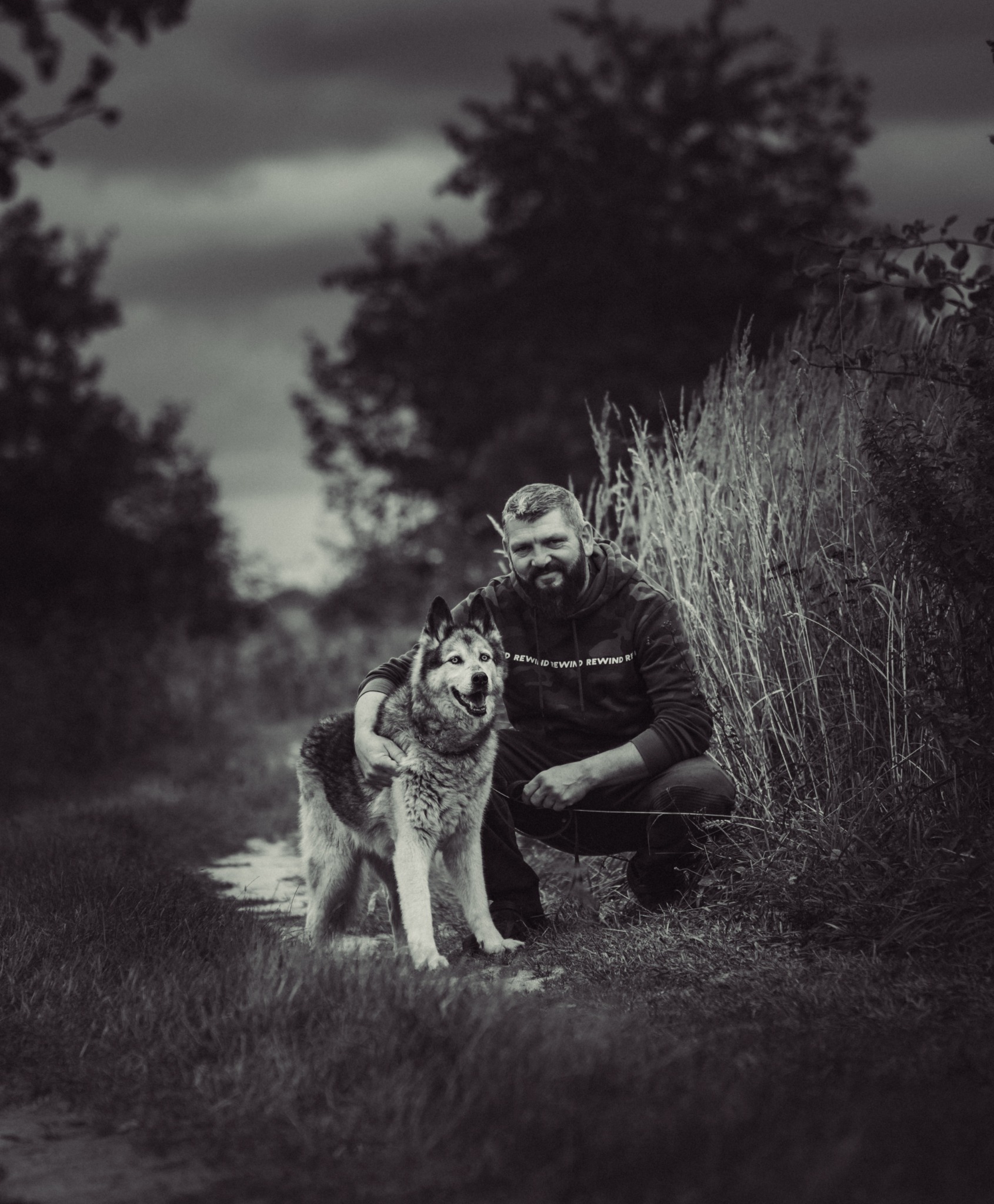 Czarno-białe zdjęcie mężczyzny z brodą, w bluzie z kapturem, kucającego obok uśmiechniętego psa rasy husky na tle łąki i pochmurnego nieba.