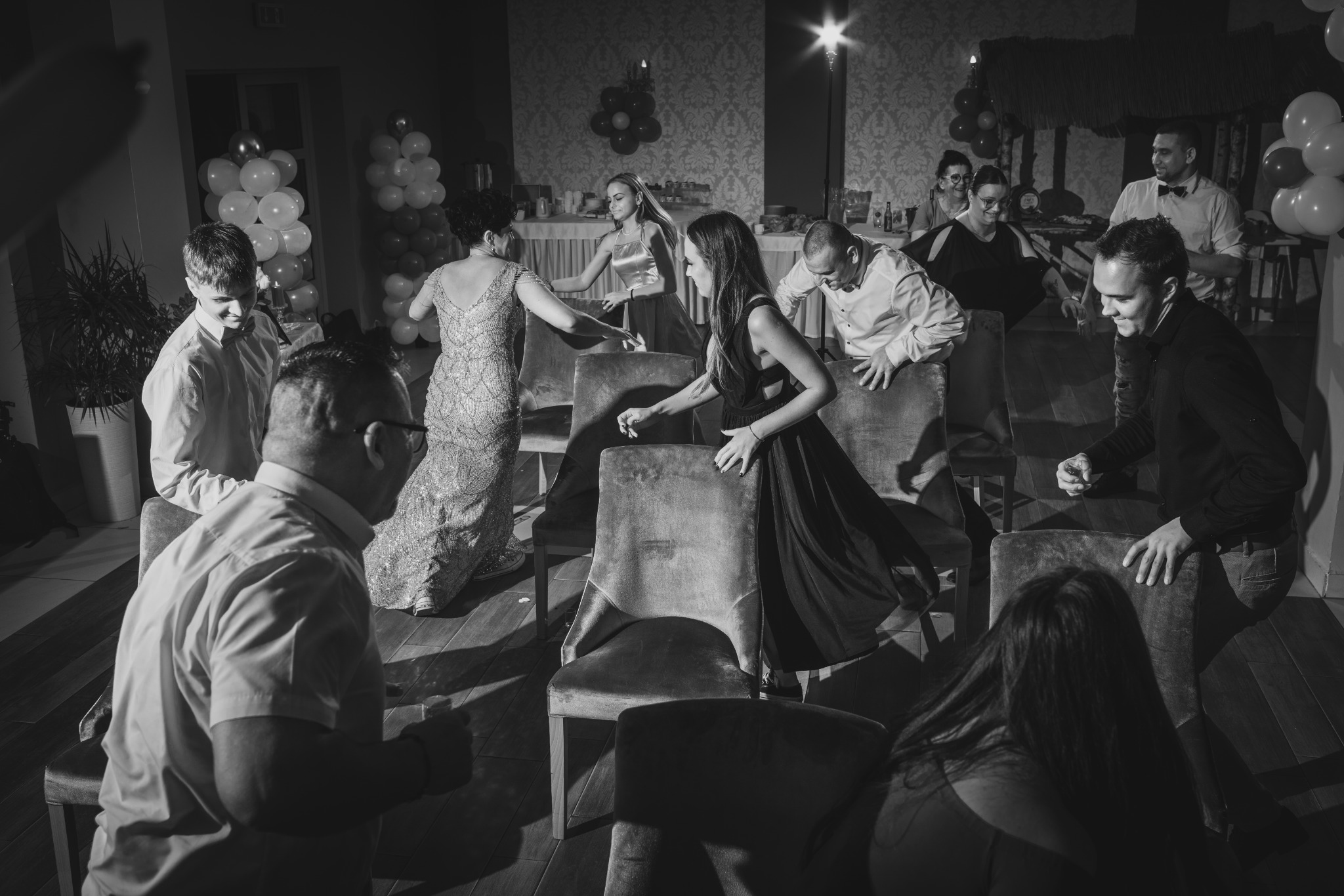 Czarno-białe zdjęcie grupy osób grających w grę siedząc na krzesłach podczas imprezy z dekoracjami balonowymi w tle.