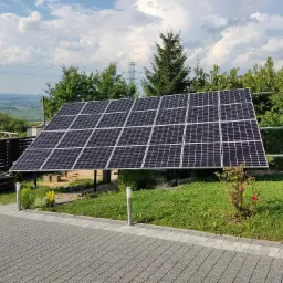 Panele słoneczne zamontowane na metalowej konstrukcji na trawniku, w tle drzewa, słupy wysokiego napięcia i błękitne niebo z chmurami.