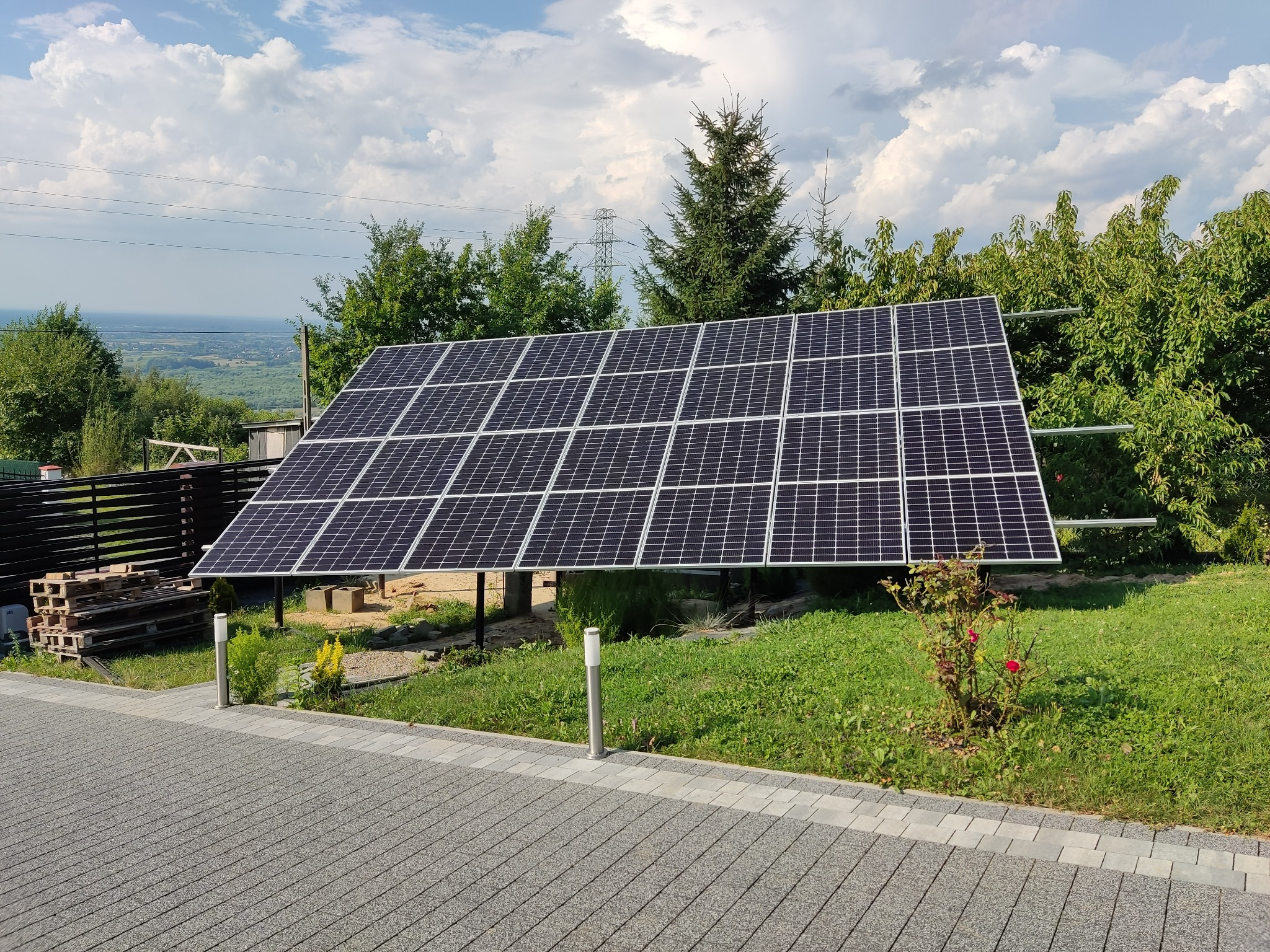 Panele słoneczne zamontowane na metalowej konstrukcji na trawniku, w tle drzewa, słupy wysokiego napięcia i błękitne niebo z chmurami.
