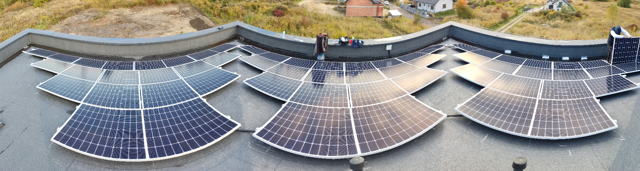 Szeroki kadr na instalację paneli słonecznych ułożonych w łuk na płaskim dachu z widoczną ekipą montażową i otoczeniem budynków mieszkalnych oraz drzew