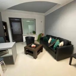 Wnętrze gabinetu terapeutycznego z czarną skórzaną sofą, stolikiem kawowym z pudełkiem chusteczek, biurkiem i krzesłami, utrzymane w stonowanej kolorystyce szarości i bieli.