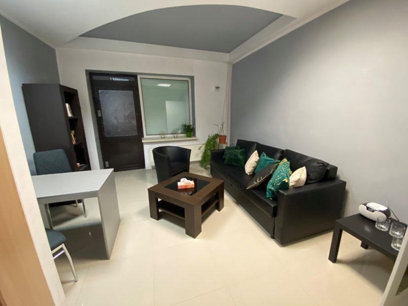 Wnętrze gabinetu terapeutycznego z czarną skórzaną sofą, stolikiem kawowym z pudełkiem chusteczek, biurkiem i krzesłami, utrzymane w stonowanej kolorystyce szarości i bieli.