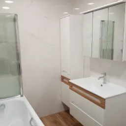 Nowoczesna łazienka z białymi płytkami imitującymi marmur, biała zabudowa umywalkowa z drewnianymi akcentami, kabina prysznicowa ze szklanymi drzwiami i lustrzana szafka nad umywalką.