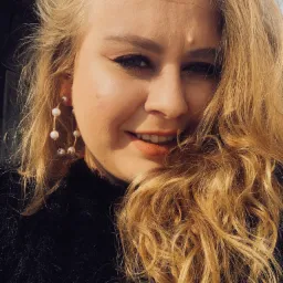 Uśmiechnięta blond kobieta w czarnym swetrze z perłowymi kolczykami, selfie w naturalnym świetle.