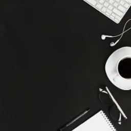 Widok z góry na minimalistyczne biurko z czarnym tłem, na którym leżą: biała klawiatura, słuchawki, filiżanka czarnej kawy, okulary, notatnik i długopis.