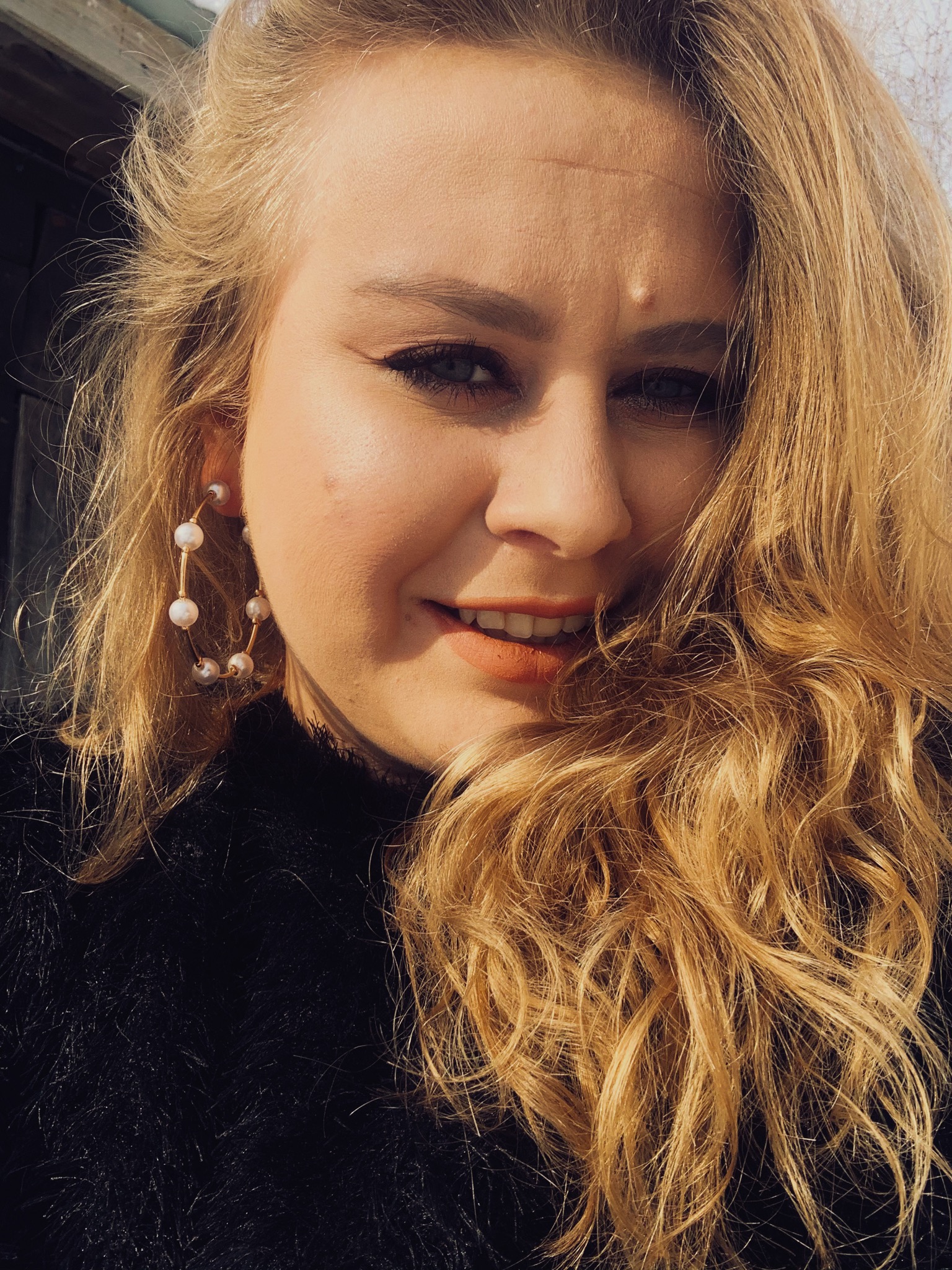 Uśmiechnięta blond kobieta w czarnym swetrze z perłowymi kolczykami, selfie w naturalnym świetle.