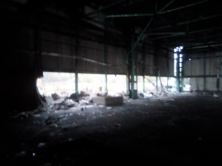 Wnętrze zdemolowanej hali przemysłowej z częściowo wyburzoną ścianą, widoczne elementy konstrukcyjne i gruz na podłodze.