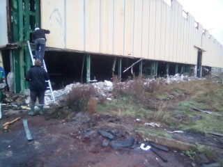 Demontaż dolnej części ściany hali przemysłowej z użyciem drabiny, widoczne elementy konstrukcyjne i gruz.