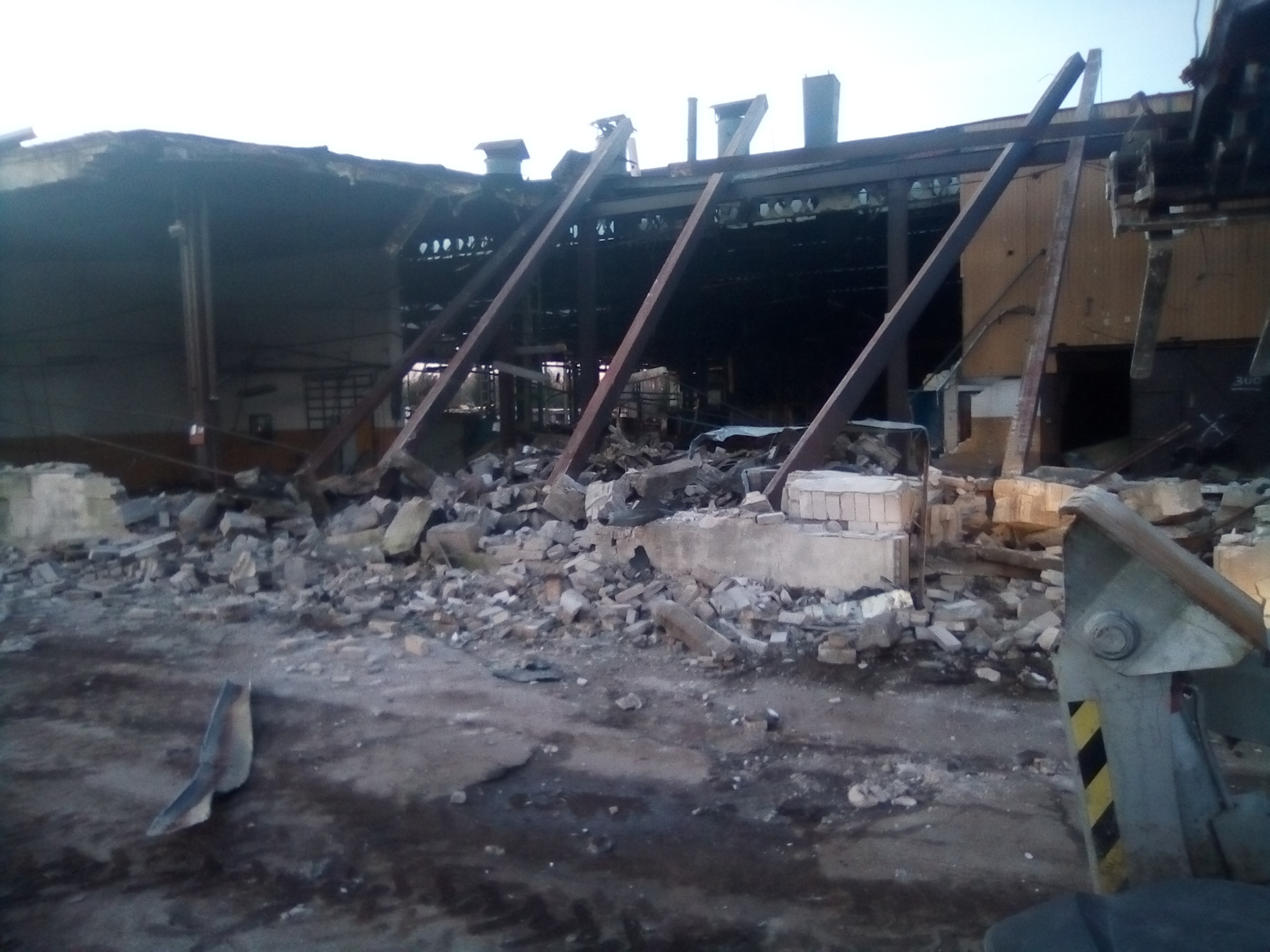 Częściowo zburzony budynek przemysłowy z widocznymi elementami konstrukcyjnymi i stertą gruzu, fragment maszyny budowlanej w prawym dolnym rogu kadru.