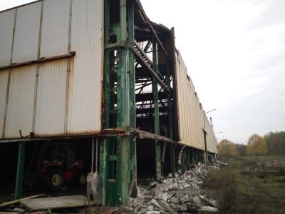 Częściowo zdemontowana hala przemysłowa z widoczną konstrukcją stalową, gruzem i wózkiem widłowym wewnątrz.