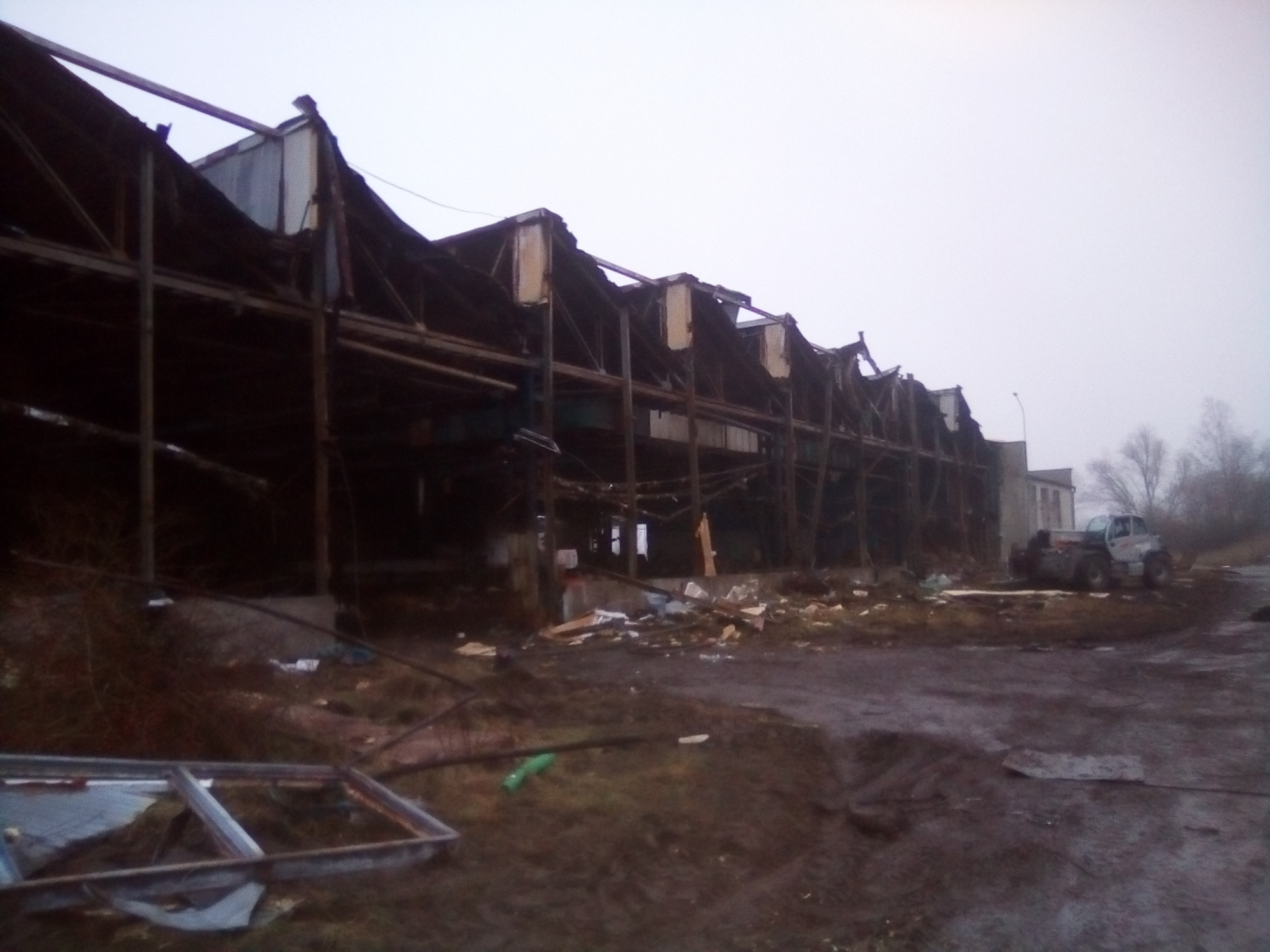 Uszkodzona konstrukcja hali przemysłowej po częściowej rozbiórce, widoczny pojazd budowlany w tle, teren błotnisty.