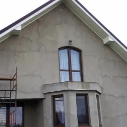 Surowa elewacja domu jednorodzinnego w trakcie prac, widoczne rusztowanie, okna z brązowymi ramami i wystający balkon, ściany w szarym tynku przed nałożeniem warstwy wykończeniowej.