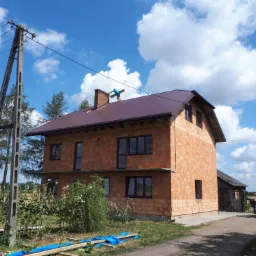 Pracownik na dachu dwupiętrowego domu z czerwonej cegły, montuje element przy kominie na tle błękitnego nieba z obłokami. Dach pokryty blachą w kolorze ciemnego brązu.