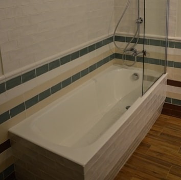 Biała wanna z parawanem nawannowym, obudowana płytkami w odcieniach beżu i bieli, z dekoracyjnym pasem zieleni, na podłodze drewnopodobne panele.