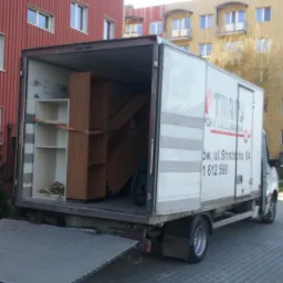 Ciężarówka transportowa z otwartą paką, załadowana meblami zabezpieczonymi pasami, na tle budynków mieszkalnych.