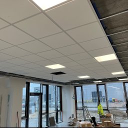 KSA SP. z o.o. - Montaż sufitu podwieszanego kasetonowego w nowym biurze, widoczne instalacje nad sufitem, duże okna z widokiem na plac budowy, w tle ubrany w odblaskową kamizelkę pracownik.