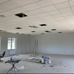 KSA SP. z o.o. - Wnętrze dużego pomieszczenia w trakcie prac wykończeniowych; widoczny biały, kasetonowy sufit podwieszany z otworami na instalacje, białe ściany, okna, grzejniki, drabina, farby, stolik...