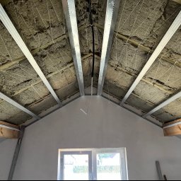 KSA SP. z o.o. - Poddasze w trakcie ocieplania: widoczne krokwie z izolacją, metalowe profile pod konstrukcję sufitu i wystające przewody elektryczne.