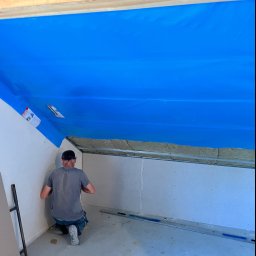 KSA SP. z o.o. - Pracownik montuje płytę karton gips na poddaszu, widoczne ocieplenie, niebieska folia paroizolacyjna, profil stalowy i młotek na podłodze.