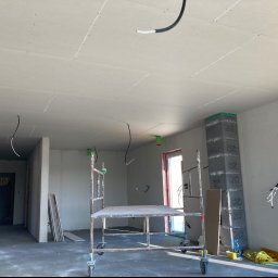 KSA SP. z o.o. - Wnętrze w surowym stanie podczas prac wykończeniowych: zamontowane płyty na suficie, widoczne kable elektryczne, rusztowanie na kółkach, mur z bloczków betonowych, okno z widokiem na zewnątrz,...