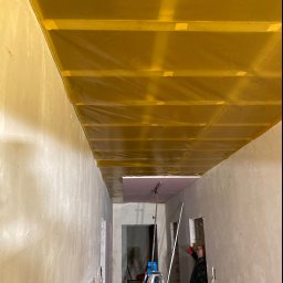 KSA SP. z o.o. - Wnętrze w trakcie remontu: żółta folia paroizolacyjna zamontowana na suficie podwieszanym, ściany w surowym tynku, drabina i pracownik w oddali, przygotowujący się do dalszych prac.