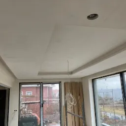 Wykończony sufit wielopoziomowy z widocznym otworem na oświetlenie, listwą dekoracyjną przy ścianie i surowymi ścianami w nowym budynku z dużymi oknami.