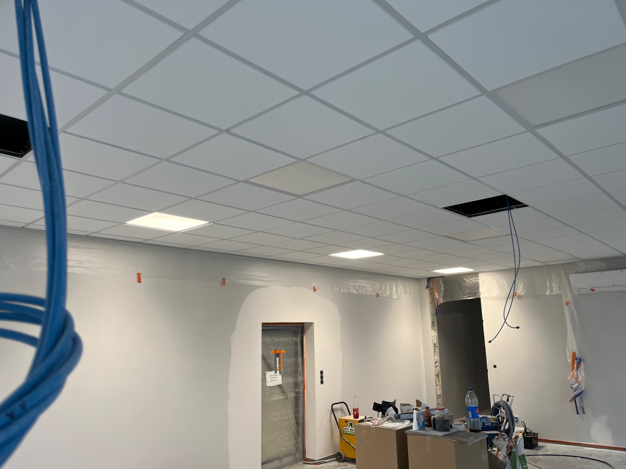 Montaż sufitu podwieszanego kasetonowego z widocznymi otworami na oświetlenie i kable, ściany przygotowane do malowania, zabezpieczone folią malarską, widoczne materiały i narzędzia pracy.