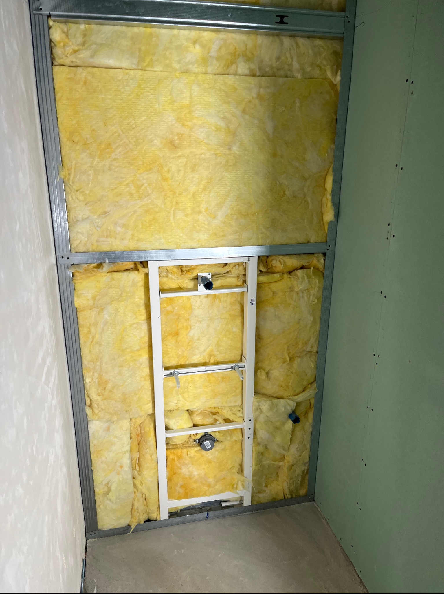 Wnętrze w trakcie prac wykończeniowych: wełna mineralna w metalowym stelażu, widoczna instalacja sanitarna i fragment ściany z płyty gipsowo-kartonowej.