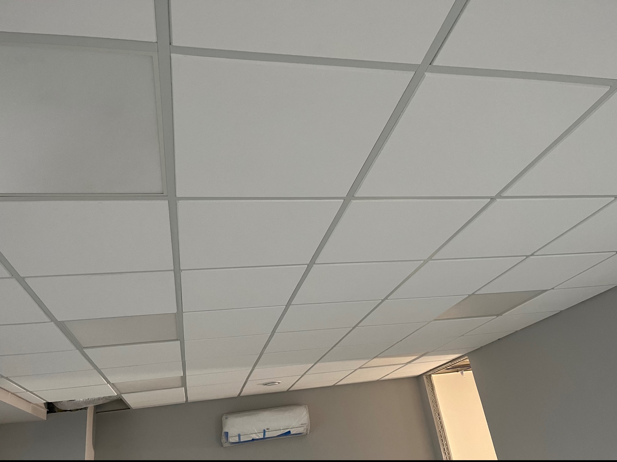 Biały sufit kasetonowy z widoczną kratownicą, kilka paneli w odcieniu szarości, klimatyzator na ścianie w folii ochronnej, widok z dołu pod kątem.