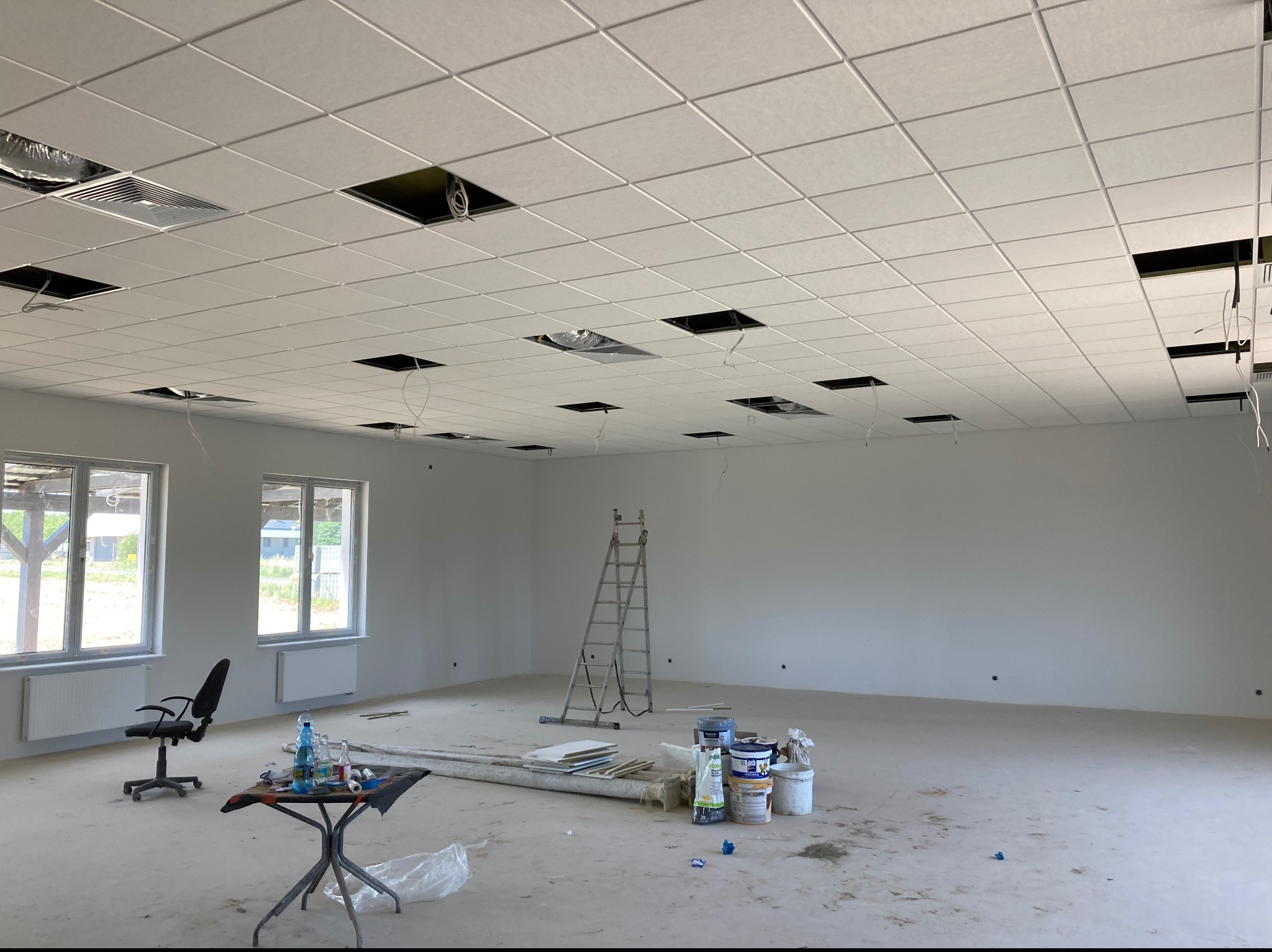 Wnętrze dużego pomieszczenia w trakcie prac wykończeniowych; widoczny biały, kasetonowy sufit podwieszany z otworami na instalacje, białe ściany, okna, grzejniki, drabina, farby, stolik...