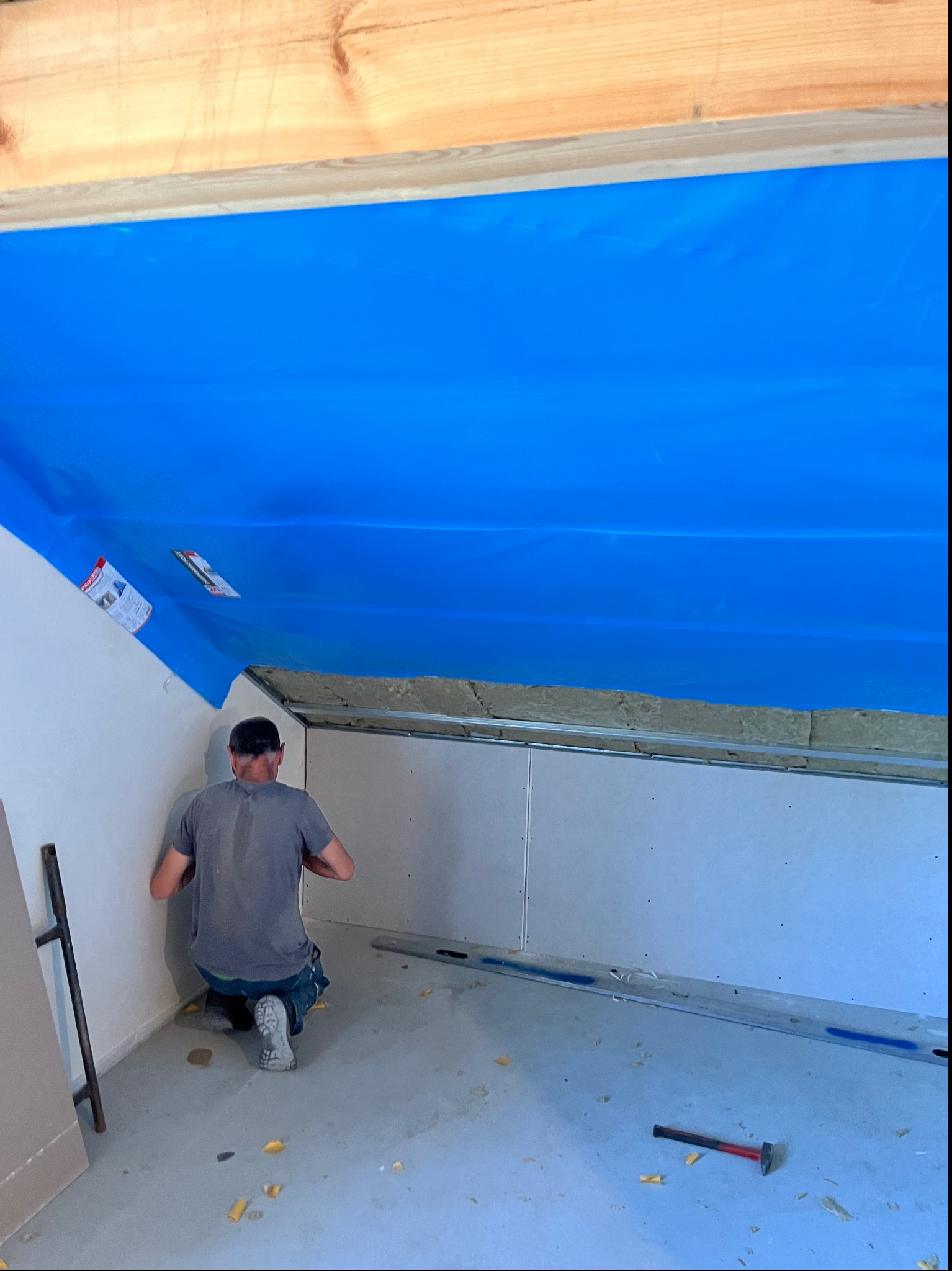 Pracownik montuje płytę karton gips na poddaszu, widoczne ocieplenie, niebieska folia paroizolacyjna, profil stalowy i młotek na podłodze.