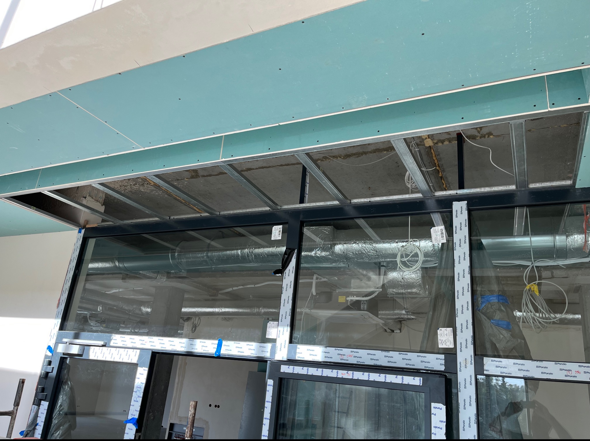 Montaż sufitu podwieszanego z płyt kartonowo-gipsowych na metalowej konstrukcji, widoczne instalacje wentylacyjne i elektryczne nad sufitem, duże okna z czarnymi ramami w folii ochronnej.