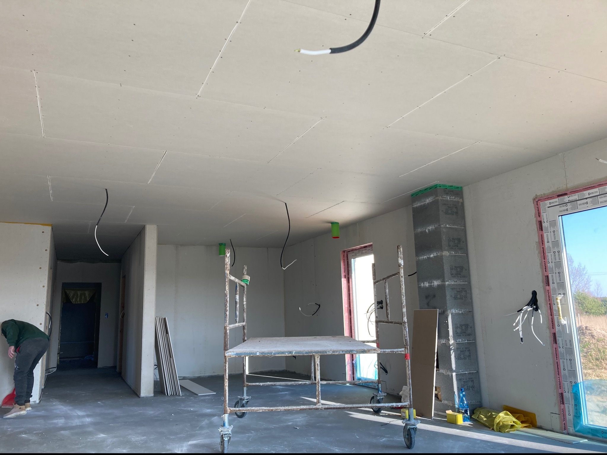 Wnętrze w surowym stanie podczas prac wykończeniowych: zamontowane płyty na suficie, widoczne kable elektryczne, rusztowanie na kółkach, mur z bloczków betonowych, okno z widokiem na zewnątrz,...