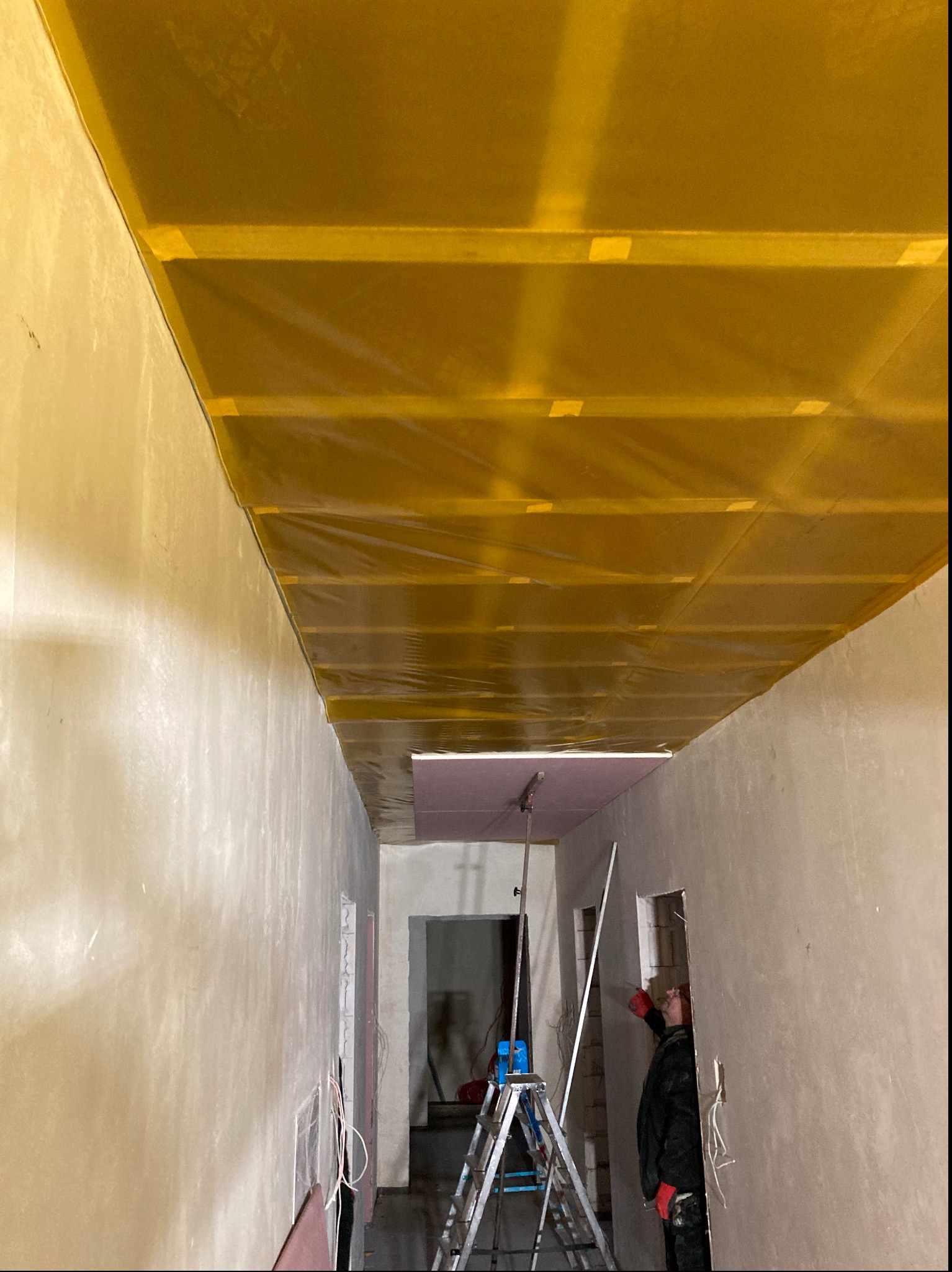 Wnętrze w trakcie remontu: żółta folia paroizolacyjna zamontowana na suficie podwieszanym, ściany w surowym tynku, drabina i pracownik w oddali, przygotowujący się do dalszych prac.