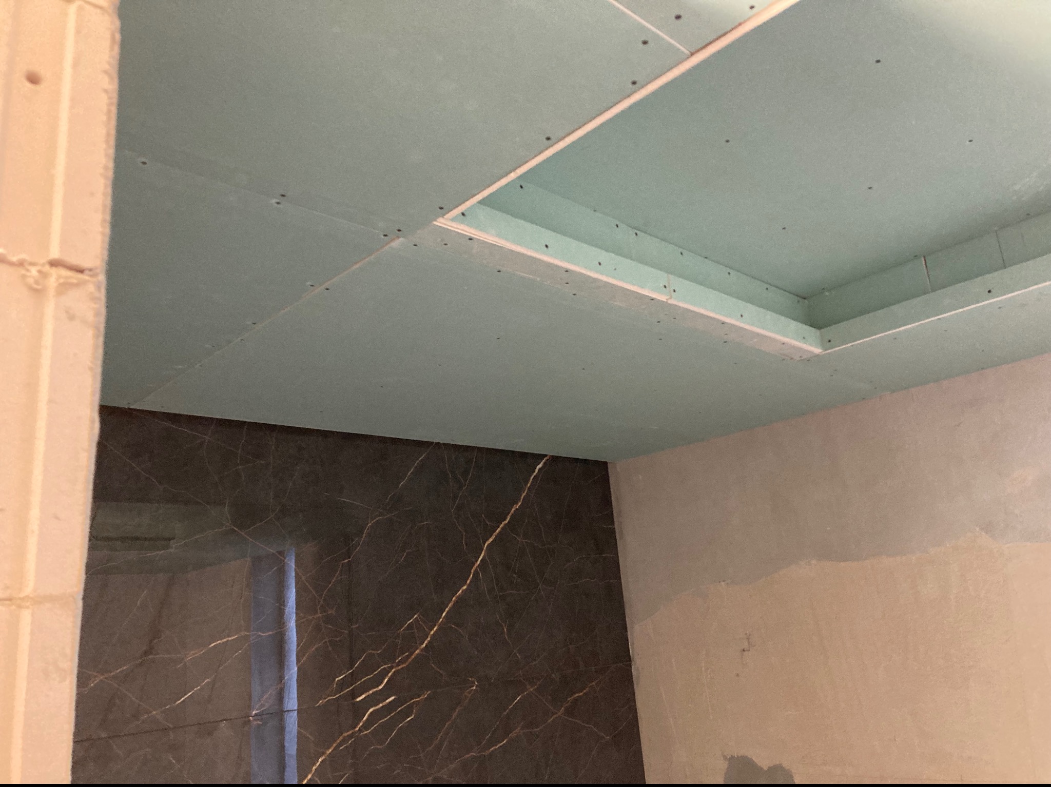 Montaż sufitu podwieszanego z płyty gipsowo-kartonowej, widoczne łączenia płyt i otwór instalacyjny, ściana wykończona płytami imitującymi marmur, surowy stan wykończenia.