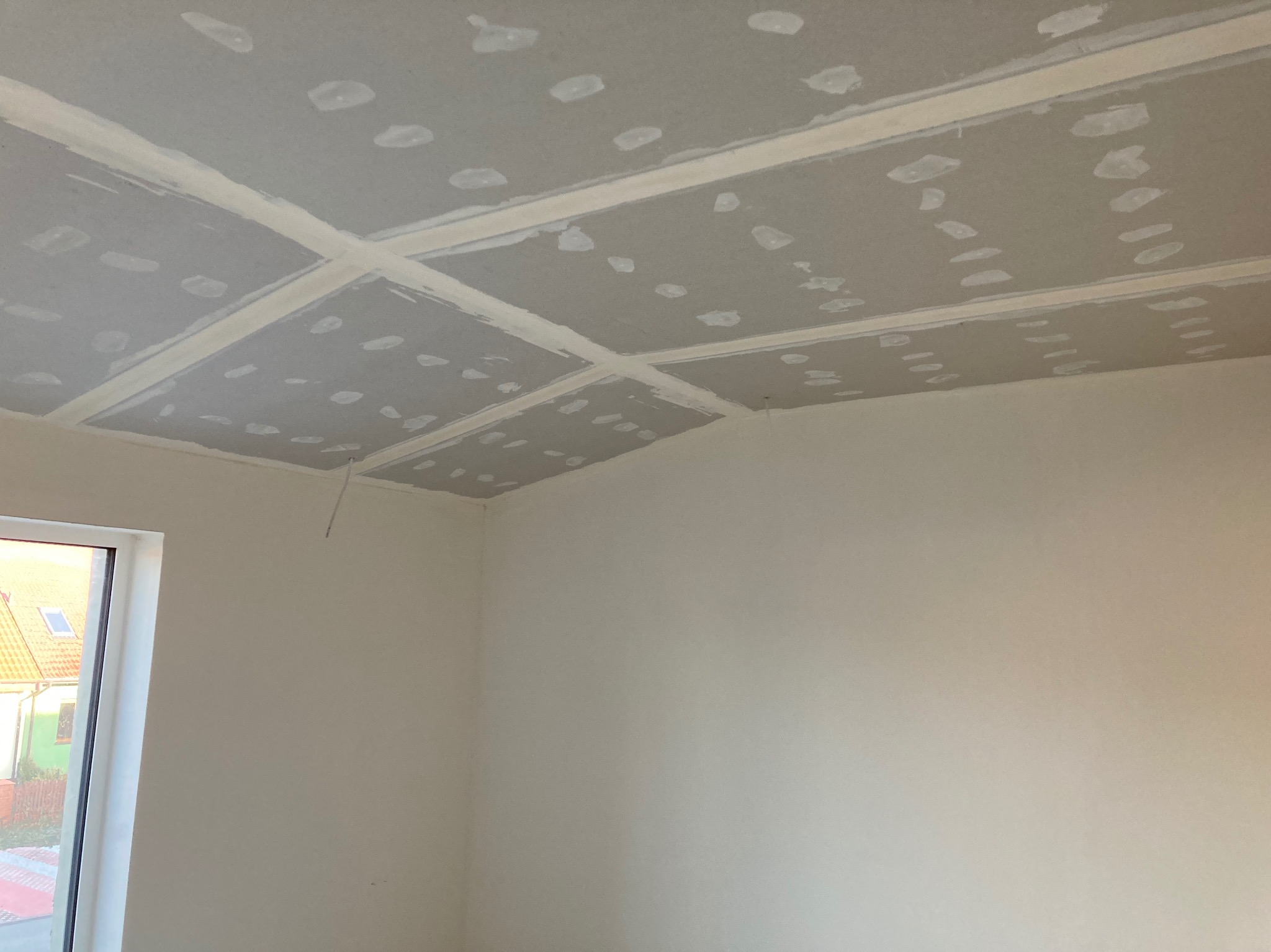 Szpachlowana i zagruntowana powierzchnia sufitu z płyt gipsowo-kartonowych, widoczne łączenia płyt wypełnione masą szpachlową, w tle okno z widokiem na dom.