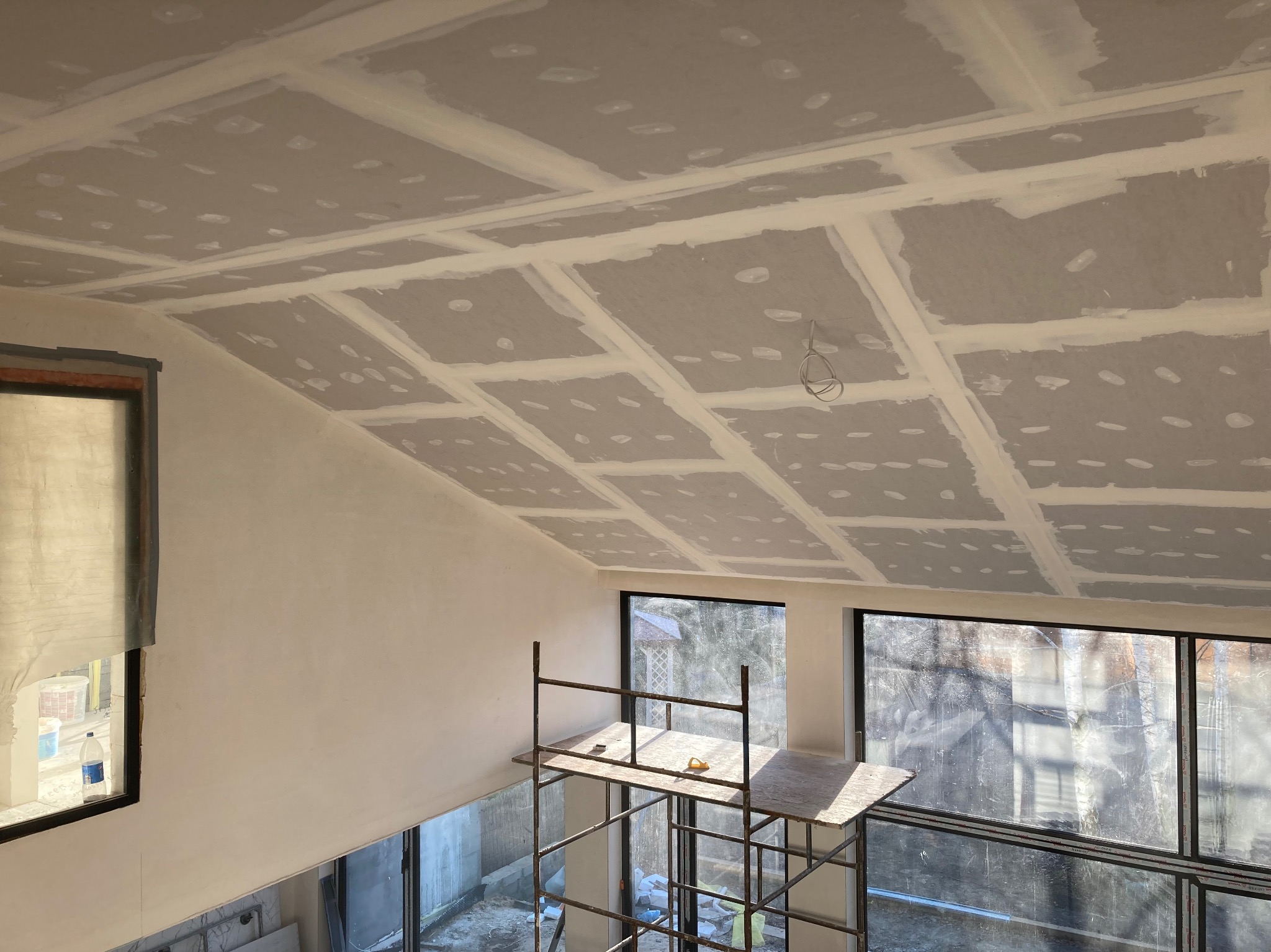 Skośny sufit poddasza pokryty płytami gipsowo-kartonowymi, widoczne łączenia szpachlowane, rusztowanie robocze pod oknem, przewody elektryczne zwisające z sufitu.