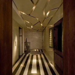 Nowoczesny korytarz z geometrycznym wzorem na suficie, wykonanym z podświetlanych listew i czarno-białą podłogą w pasy. Drewniane drzwi po prawej stronie, metalowa rzeźba w tle.