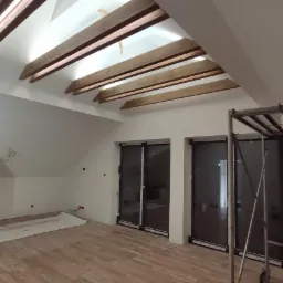 Poddasze w trakcie wykańczania, widoczne drewniane belki stropowe, ściany przygotowane do malowania, okna przesuwne i rusztowanie malarskie.