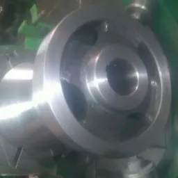 Metalowy element z otworami zamocowany w uchwycie tokarki, widoczna część zielonej obudowy maszyny.