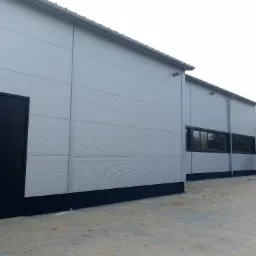 Szary budynek przemysłowy z panelami, czarnymi oknami i drzwiami, oraz widocznym systemem rynnowym.