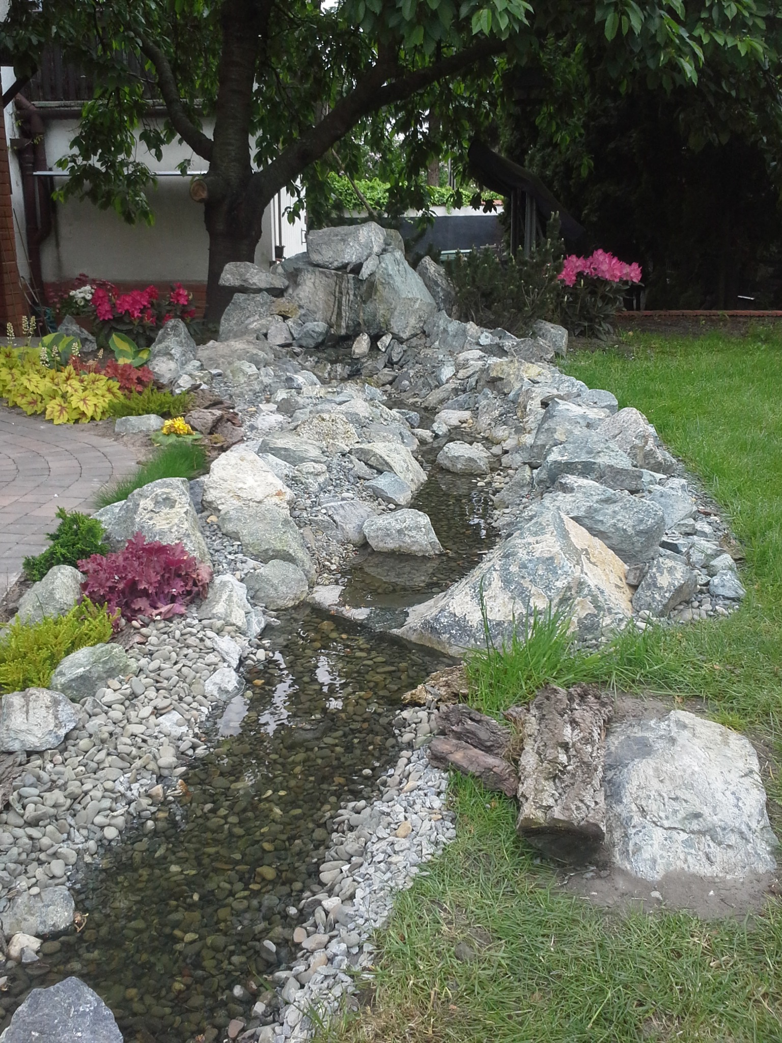 Aranżacja ogrodu z kaskadą wodną zbudowaną z kamieni, otoczoną roślinami i trawą, z drzewem w tle.