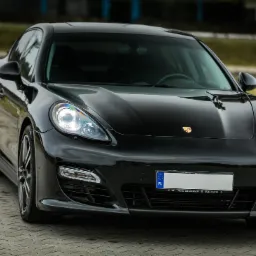 Błyszczący, czarny samochód osobowy Porsche Panamera z włączonymi światłami mijania, zaparkowany na kostce brukowej, widok z przodu na zewnątrz.