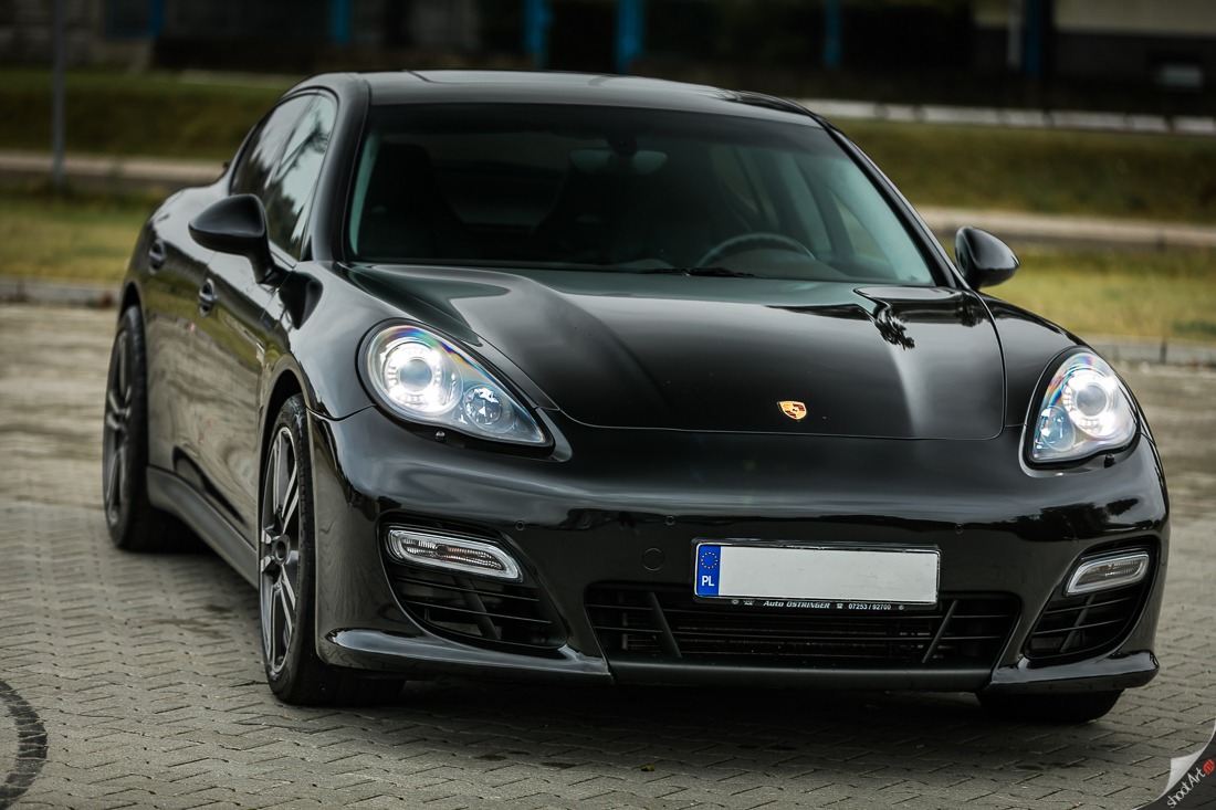 Błyszczący, czarny samochód osobowy Porsche Panamera z włączonymi światłami mijania, zaparkowany na kostce brukowej, widok z przodu na zewnątrz.