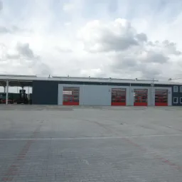 Widok na budynek z trzema czerwonymi bramami garażowymi i zadaszoną wiatą, pod którą stoją wózki widłowe, na pierwszym planie szara nawierzchnia z kostki brukowej.