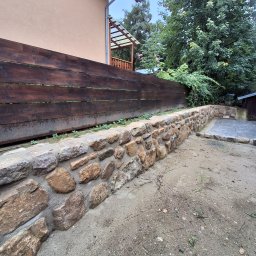 Firma Brukarska Roboty Ziemne Dariusz Karykkowski - Kamienny murek oporowy z drewnianym ogrodzeniem w tle, otoczony zielenią. Ujęcie z perspektywy ziemi ukazuje solidną konstrukcję i naturalne materiały.
